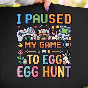 May include: Black t-shirt with a colorful graphic that reads "I PAUSED MY GAME TO EGG HUNT." The design includes a game controller, Easter eggs, a basket, a handheld game console, and bunnies.