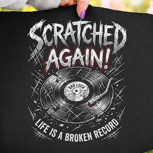 Könnte beinhalten: Schwarzes T-Shirt mit einem verwitterten Grafikdesign einer zerbrochenen Schallplatte. Der Text lautet "Scratched Again!" und "Life is a Broken Record." Das Design beinhaltet eine Plattenspielernadel und die Worte "Bad Luck."