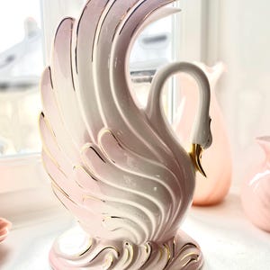 Op de afbeelding: Een roze en witte keramische zwaanfiguur met gouden details. De zwaan heeft een gebogen nek en gedetailleerde vleugelveren. De basis en randen zijn afgewerkt met goud. Een decoratief item voor in huis.