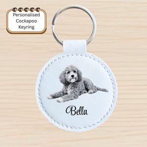 May include: A personalized Cockapoo keyring featuring a light blue circular design with a grayscale image of a Cockapoo dog named Bella. The keyring has a silver metal ring and a white stitched border.