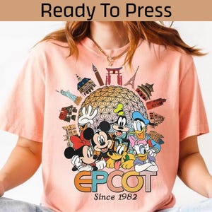 May include: A pale pink t-shirt with a graphic of Disney characters, including Mickey and Minnie Mouse, Goofy, Donald and Daisy Duck. The shirt has the text "EPCOT Since 1982" and the words "Ready To Press" at the top.