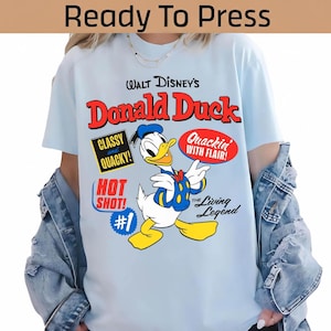 Può includere: T-shirt azzurra con grafica di Paperino e la scritta "WALT DISNEY'S Donald Duck". Il design include le frasi "Classy and Quacky!", "Hot Shot! #1", "Quackin' with Flair!" e "The Living Legend".