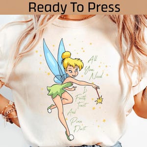 May include: Cream-colored t-shirt featuring a cartoon illustration of a fairy with yellow hair, green dress, and blue wings. The text "All You Need Is Faith, Trust, And Pixie Dust" is also printed on the shirt.