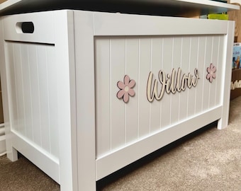 May include: A white wooden toy chest with a hinged lid and side handles. The front panel features vertical slats, the name Willow in cursive script, and two light pink flower cutouts on either side of the name, all mounted on the white surface.