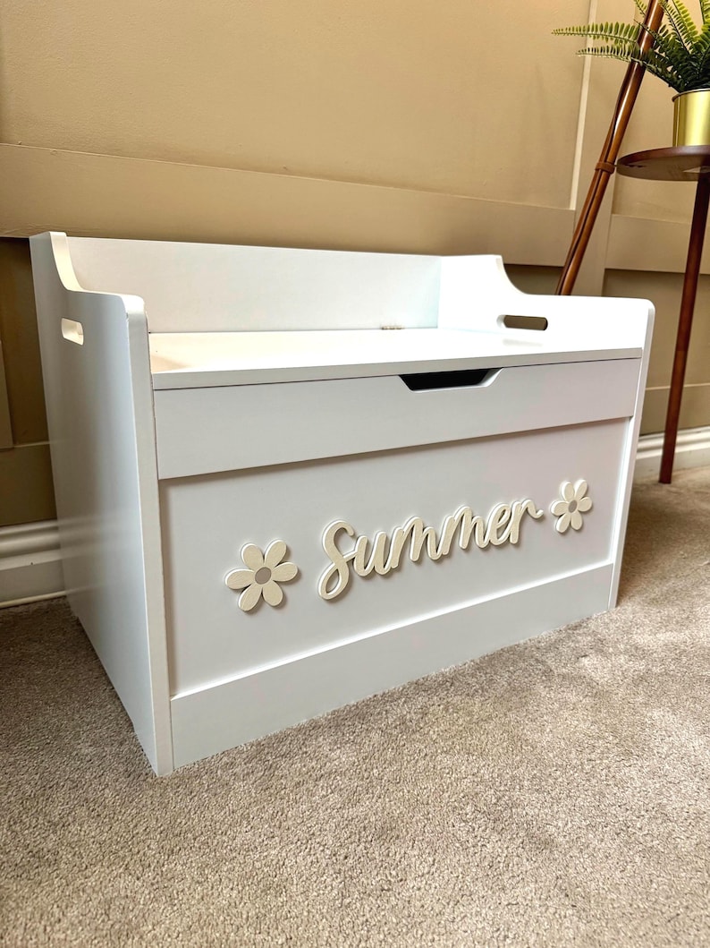 May include: White wooden toy chest with a hinged lid and a front panel featuring the word "Summer" in cursive script, accompanied by two flower accents. The chest has a handle on the front and cut-out handles on the sides.