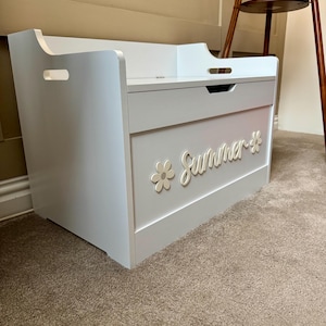 May include: A white and gray wooden toy chest with the word "Summer" and a flower design on the front. The chest has a lid with a handle and cut-out handles on the sides. The chest is sitting on a brown carpet.