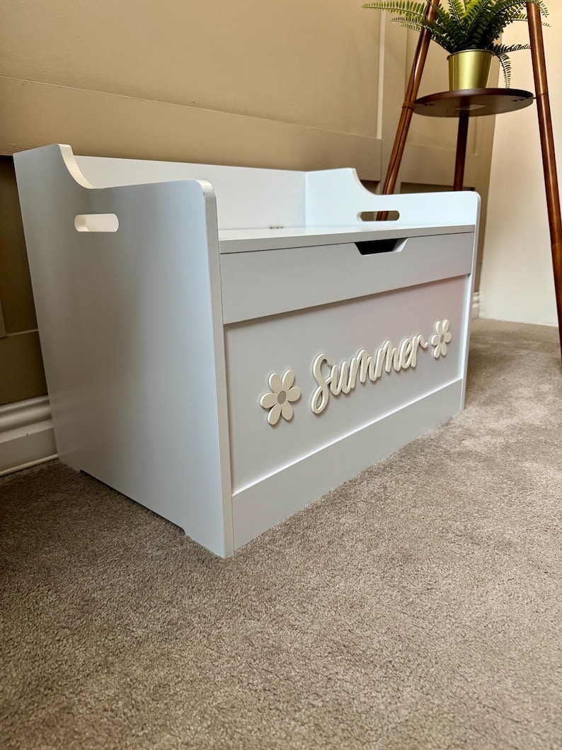 May include: A white and gray wooden toy chest with the word "Summer" and a flower design on the front. The chest has a lid with a handle and cut-out handles on the sides. The chest is sitting on a brown carpet.
