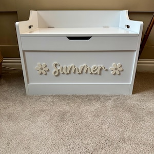 May include: A white wooden toy chest with a hinged lid and a handle. The word "Summer" is spelled out in cursive with two flower accents. The chest is sitting on a beige carpet.