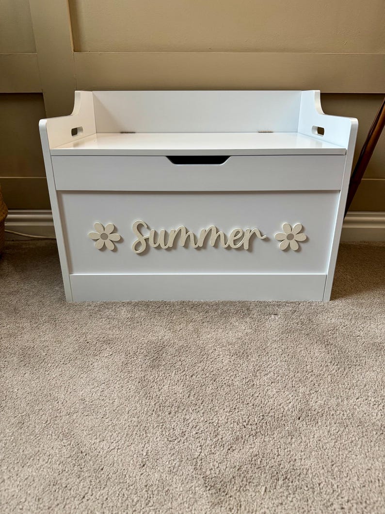 May include: A white wooden toy chest with a hinged lid and a handle. The word "Summer" is spelled out in cursive with two flower accents. The chest is sitting on a beige carpet.