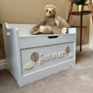 May include: A pale grey wooden toy chest with the word "Summer" in cursive, hot air balloon and cloud decorations. A plush sloth toy sits on top. The chest has a bench-like top and cut-out handles.