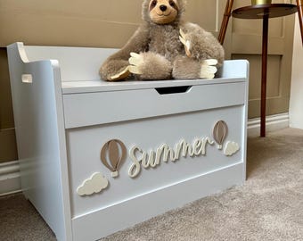 May include: A light gray wooden toy chest with the word "Summer" in cursive, along with hot air balloon and cloud accents. A plush sloth toy sits on top. The chest has a bench-like top and cut-out handles.