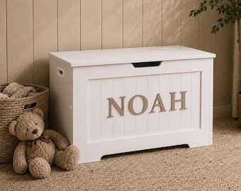 May include: A white rectangular toy chest with a hinged lid and a cutout handle. The front panel features vertical slats and the name NOAH in large, raised, light brown capital letters. The chest has side handles and a smooth, clean finish.