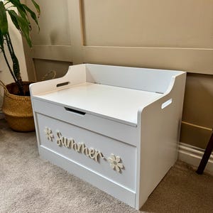 May include: A white wooden toy chest with a flat top and cut-out handles. The word "Summer" is displayed on the front in cursive script, flanked by flower accents. The chest is placed on a carpeted floor.