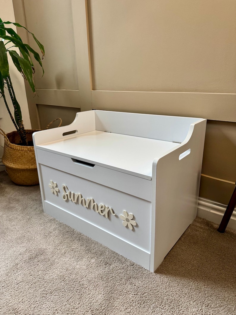 May include: A white wooden toy chest with a flat top and cut-out handles. The word "Summer" is displayed on the front in cursive script, flanked by flower accents. The chest is placed on a carpeted floor.