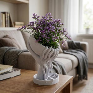 Könnte beinhalten: Ein dekorativer Pflanzentopf in Form von Händen, die einen Strauß lila Blumen halten. Der Topf ist hellgrau und hat eine strukturierte Oberfläche. Die Blumen sind ein leuchtendes Lila. Der Topf steht auf einem runden Sockel.
