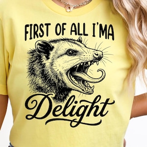 Könnte beinhalten: Gelbes T-Shirt mit einer schwarz-weißen Opossum-Grafik und dem Text "FIRST OF ALL I'M A Delight". Das Opossum wird mit offenem Mund und herausgestreckter Zunge dargestellt. Das Shirt besteht aus einem weichen Material.