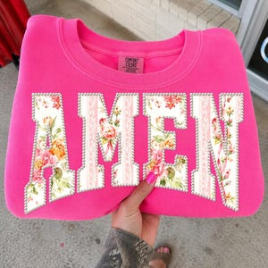 Op de afbeelding: Een felroze sweatshirt met het woord "AMEN" in grote letters met een bloemenpatroon. De letters zijn omrand met kleine, zilverkleurige kralen. De sweatshirt heeft een ronde hals en het merk "Comfort Colors" is zichtbaar.