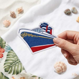 May include: An embroidered patch shaped like a cruise ship with a dark blue hull, a red bottom, and white upper decks. The ship features two red smokestacks, gold decorative scrollwork on the bow, and a white border outlining the entire shape.