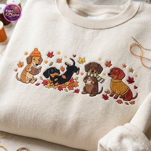 May include: A beige crewneck sweatshirt featuring an embroidered design of four dachshunds in an autumn setting. The dogs are depicted reading a book, playing in fallen leaves, holding a warm mug, and wearing a yellow sweater, surrounded by leaves.