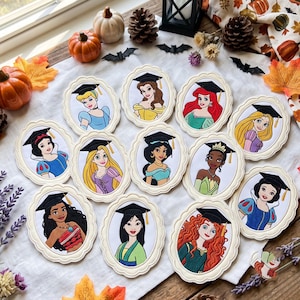 May include: A collection of twelve oval-shaped embroidered patches featuring various animated princesses wearing black graduation caps with yellow tassels. Each patch is framed in a scalloped cream-colored border, showcasing detailed character portraits.