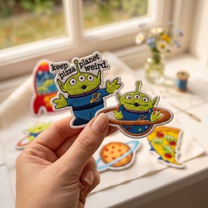 Può includere: Due toppe ricamate raffiguranti alieni verdi a tre occhi in tute spaziali blu. Una toppa mostra un alieno con le braccia tese e il testo "keep pizza planet weird". L'altra mostra un alieno all'interno di un pianeta marrone con anelli.