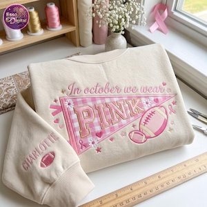 Op de afbeelding: Een crèmekleurig sweatshirt met borduurwerk met de tekst In october we wear PINK. Het ontwerp bevat een roze gingham wimpel, een football, madeliefjes, sterren, hartjes en linten. Op de mouw is de naam CHARLOTTE geborduurd naast een football.