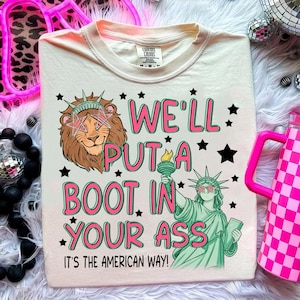 May include: Cream-colored t-shirt with a graphic design featuring a lion wearing star-shaped sunglasses and the Statue of Liberty. The text reads "WE'LL PUT A BOOT IN YOUR ASS. IT'S THE AMERICAN WAY!" in pink and black.