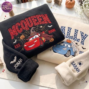 May include: A black sweatshirt featuring an embroidered red race car with a witch hat and spiderweb, text reading MCQUEEN EST. 2006, and a sleeve with a bat and the name JONAH. Beside it, a cream sweatshirt with a blue car, spiderweb, and name SARAH.
