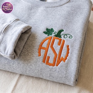 Pode incluir: Uma camisola de gola redonda cinzento-claro com um desenho bordado no peito. O bordado representa uma abóbora cor de laranja formada pelas letras ASW, rematada com um caule verde e uma folha verde. O tecido tem uma textura mesclada.