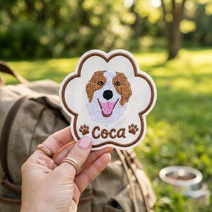 May include: A paw-shaped embroidered patch featuring a brown and white dog portrait with a pink tongue, the name "Coca" in brown thread, and two small brown paw prints on a cream-coloured fabric background with a thick brown stitched border.