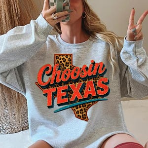 Może przedstawiać: Jasnoszara bluza z napisem "Choosin TEXAS" w stylu retro. Tekst jest czerwony i niebieski, z obrysem Teksasu w panterkę. Bluza ma swobodny styl.