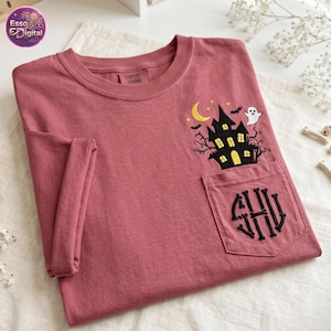 Pode incluir: Uma t-shirt de manga curta em rosa seco com uma casa assombrada preta bordada, uma lua crescente amarela, morcegos e um fantasma branco. O bolso no peito apresenta um monograma circular feito de ossos pretos com as letras SHV.