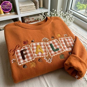 Puede incluir: Una sudadera de color naranja quemado con la palabra FALL bordada en letras grandes. Cada letra está sobre un parche cuadrado de cuadros vichy blancos y naranjas. El diseño incluye bellotas, hojas y pequeñas flores blancas bordadas.