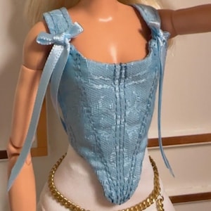 Puede incluir: Una muñeca con cabello rubio recogido en una coleta alta, que viste un corsé azul claro con lazos en los hombros y una falda blanca con un cinturón de cadena dorada. La muñeca presenta detalles faciales pintados, incluyendo ojos azules.