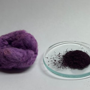 Könnte beinhalten: Nahaufnahme einer Kugel aus lila Wolle und einer kleinen Glasschale mit einem Haufen dunkelviolettem Pulver. Die Wolle ist weich und strukturiert, während das Pulver körnig erscheint.