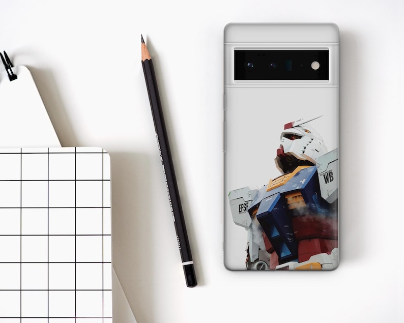 May include: A white phone case featuring a colorful illustration of a robot. The robot has a white head, red accents, and blue and yellow details. A black pencil and a notepad with a grid pattern are also visible.