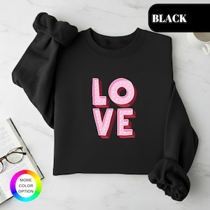 May include: Black sweatshirt with the word "LOVE" in pink, 3D-style lettering. The letters have a dotted outline and a red shadow. The sweatshirt has long sleeves and a crew neck.