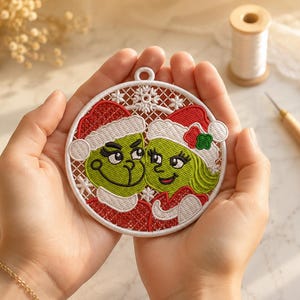 May include: A circular white embroidered ornament featuring two green characters with large eyes and eyelashes, both wearing red and white Santa hats. The design includes a white lattice background with snowflake accents and a small loop at the top.