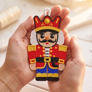 Op de afbeelding: Een notenkraker-ornament met gedetailleerd borduurwerk in rode, blauwe, gele en zwarte draad. Het ontwerp bevat een hoge rode hoed met gele accenten, een zwarte snor, wit haar en een uniform met gouden knopen en een zwarte riem.