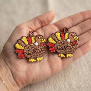 May include: Two embroidered turkey charms featuring brown bodies, yellow and red tail feathers, yellow beaks, and red wattles. Each turkey has a small circular loop at the top for hanging, with detailed stitching throughout the design.