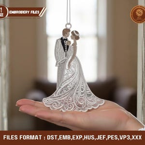 May include: An embroidered wedding couple ornament. The groom is in a tuxedo, and the bride wears a lace wedding dress. The ornament hangs from a silver string. The text "EMBROIDERY FILES" is visible.