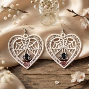 May include: A pair of white heart-shaped lace earrings featuring an intricate spider web pattern and a small black embroidered spider hanging from the center. Each earring includes a small pink heart detail at the bottom and a silver-toned metal hook.