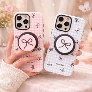 May include: Two phone cases with bow designs. One case is pink and the other is light blue, both with a gingham pattern and small bow accents. Each case has a matching pop-up grip with a bow design.