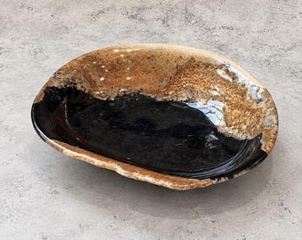 May include: An oval ceramic bowl with an irregular, organic rim. The interior features a deep, glossy black glaze at the base, transitioning to a textured, speckled tan and brown glaze along the upper walls and the outer edge of the rim.