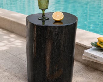 Pode incluir: Uma mesa lateral cilíndrica de madeira preta com um acabamento escuro e liso. Em cima, há um copo de vidro verde texturizado e meio limão amarelo. A mesa está posicionada ao ar livre perto de uma piscina, com um livro visível por perto.