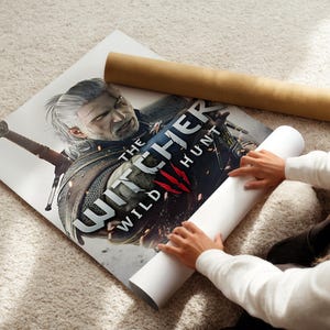 A person is holding a poster of a character from the video game The Witcher, which features a sword and a detailed illustration of the character.