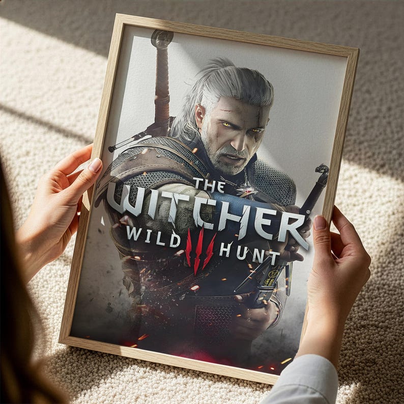 A person is holding a framed poster of a character from the video game The Witcher, featuring the character in a fighting stance.