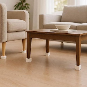 Könnte beinhalten: Ein Wohnzimmer mit einem braunen Holztisch, einer weißen Schüssel und Büchern. Zwei beige Sessel und ein Sofa befinden sich im Hintergrund. Weiße Möbelbeinschutz sind an den Beinen der Möbel angebracht.