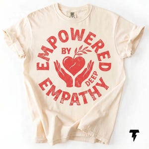 May include: Cream-colored t-shirt with a red graphic. The graphic reads "EMPOWERED BY DEEP EMPATHY" in a circular design, featuring a heart held by hands and a sprig. The shirt has rolled-up sleeves.