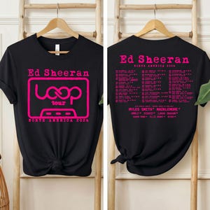 Könnte beinhalten: Ein schwarzes Kurzarm-T-Shirt mit einer pinkfarbenen Grafik einer Kassette und dem Text Ed Sheeran Loop Tour North America 2026 auf der Vorderseite sowie einer Liste von Tourdaten, Orten und Künstlernamen in Pink auf der Rückseite.